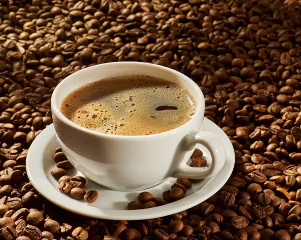 A white cup of coffee sits on a saucer surrounded by roasted coffee beans. Czy kawa rozpuszczalna podnosi ciśnienie?