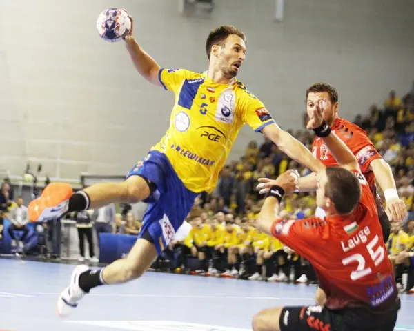 Piłkarz ręczny Telekom Veszprem w żółtej koszulce skacze do rzutu, broniony przez zawodnika w czerwonej.
