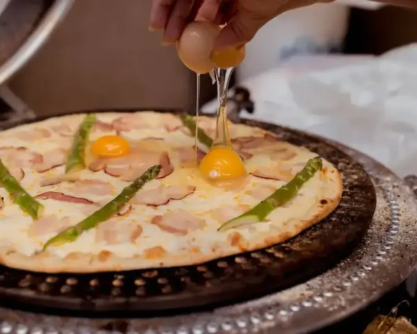 Ręka wylewa jajko na pizzę z boczkiem i szparagami. Pyszna pizza z jajkiem w przygotowaniu.