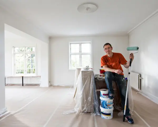 Man med färgroller och hinkar redo att måla bakom elementet i ett rum under renovering.