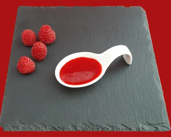 Quatre framboises fraîches à côté d'une cuillère blanche remplie de coulis de framboise, sur une ardoise.