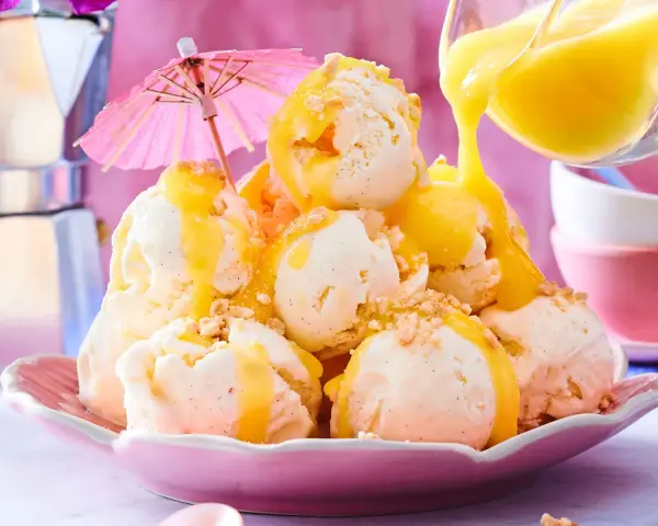 Ein Berg aus cremigem Vanilleeis, übergossen mit warmer Zitronensauce und bestreut mit Krümeln. Ein rosa Sonnenschirm sorgt für tropisches Flair.