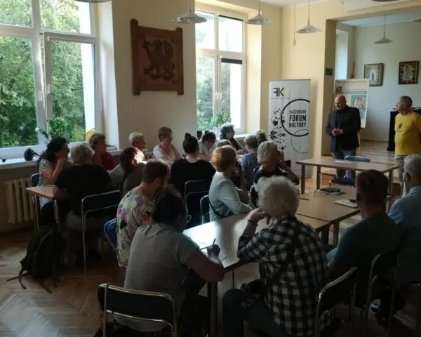 Spotkanie promujące język kaszubski. Ludzie siedzą przy stołach, słuchając prelegentów.
