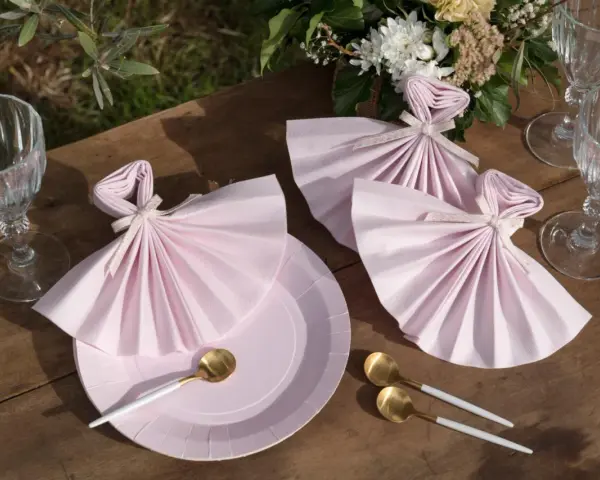 Vackert dukat bord med rosa servetter vikta som klänningar, guldfärgade skedar och glas. En bukett blommor fulländar dukningen.