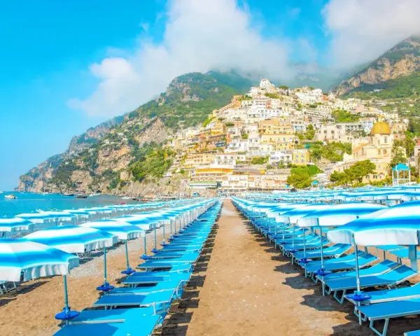Rzędy leżaków i parasoli na plaży w Positano, z widokiem na kolorowe domy wspinające się po zboczu góry. Idealne miejsce na wakacje, jak na Capri.