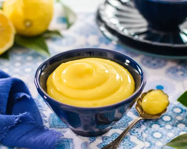 Una ciotola blu piena di vellutata crema al limone, con limoni freschi e un tovagliolo blu sullo sfondo.