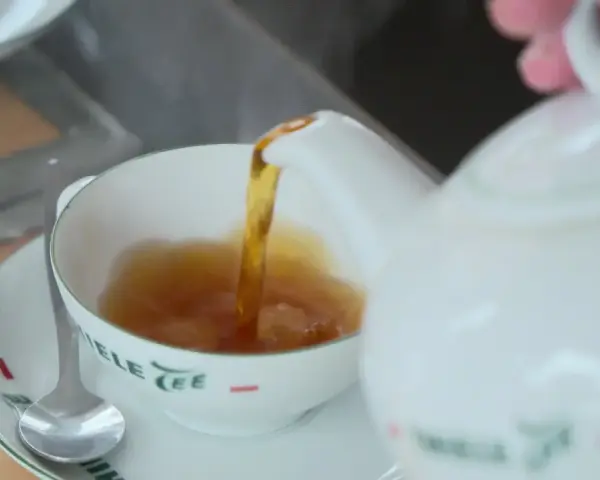 Tee mit Rum norddeutsch: Eine weiße Kanne gießt heißen Tee in eine Tasse. Ein Löffel liegt auf der Untertasse.