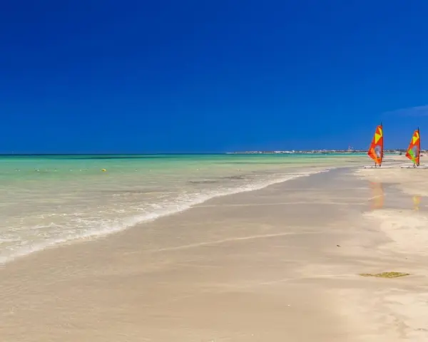 Plaża w Tunezji z trzema kolorowymi żaglami do windsurfingu na piasku i turkusową wodą.