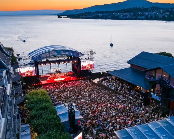 Ein großes Publikum feiert bei Sonnenuntergang ein Konzert am See. Die Bühne des Jazz Montreux Festivals ist beleuchtet, im Hintergrund der Genfersee.