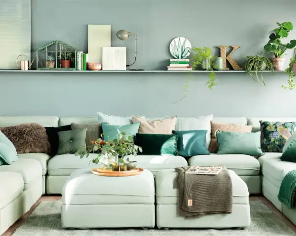 Sofá modular claro con cojines verdes y estampados, y una estantería con plantas y objetos decorativos, incluyendo una casita de cristal con **muebles pintados en verde**.