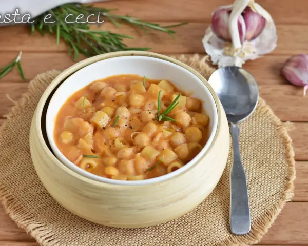 Un piatto fumante di pasta e ceci, guarnito con rosmarino fresco, su un tavolo di legno. Accanto, spicchi d'aglio e un cucchiaio.