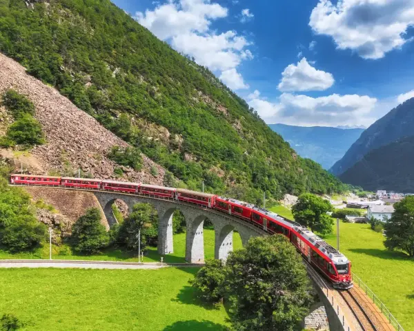 Czerwony pociąg Bernina Express przejeżdża przez kamienny wiadukt w Alpach. Sprawdź bernina express cena, by zaplanować podróż.