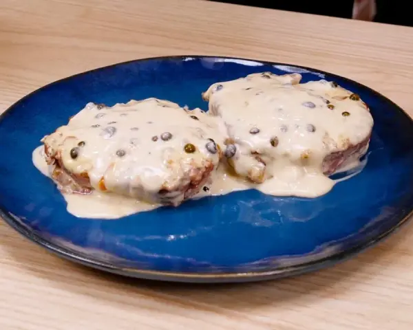 Due medaglioni di filetto al pepe verde, immersi in una cremosa salsa bianca, serviti su un piatto blu.