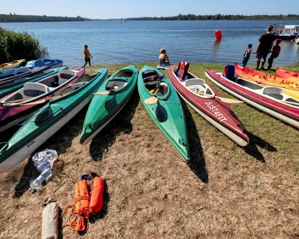 Kajaki czekają nad jeziorem. Kilka osób przygotowuje się do wodnych przygód, a w tle widać łodzie i żaglówki.