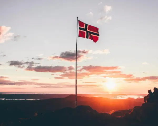 Zachód słońca w Norwegii. Dwie osoby siedzą na skale, patrząc na krajobraz. Flaga Norwegii powiewa na maszcie. Czy do Norwegii można lecieć na dowód osobisty?