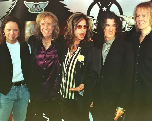 Steven Tyler mlody z zespołem Aerosmith, w charakterystycznych okularach i stroju, gotowy na scenę.