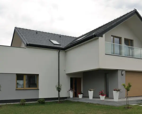 Nowoczesna elewacja domu z szarymi i białymi tynkami, dużym garażem i balkonem z przeszkloną balustradą.