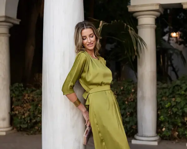 Una mujer con un vestido verde oliva satinado, como una de las madrinas elegantísimas, posa apoyada en una columna de mármol.