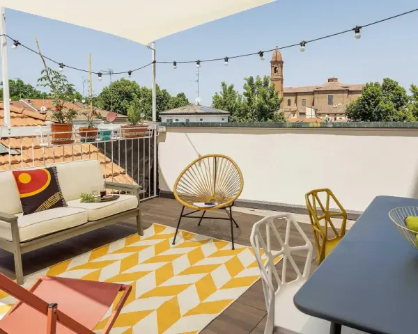 Terrazza sul tetto idee: divano, sedie colorate, tappeto giallo e bianco, tavolino con frutta, luci e vista sui tetti.
