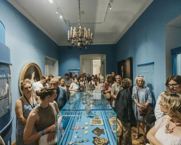 Grupa zwiedzających ogląda wystawę w muzeum. Wśród eksponatów w gablocie widać ozdobne przedmioty, a na ścianach obrazy i tablice informacyjne. Klasyczny wystrój sali z żyrandolem stanowi tło dla tego współczesnego zainteresowania historią.