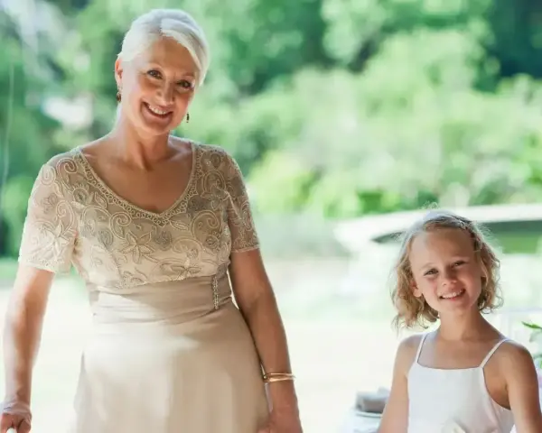 Elegancka stylizacja na komunię dla babci. Uśmiechnięta kobieta w beżowej sukni z haftem i dziewczynka w bieli.