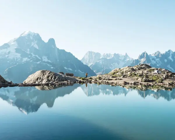 Un lac alpin reflète des montagnes majestueuses, un aperçu des plus belles randonnées de France. Une personne se tient sur la rive rocheuse.