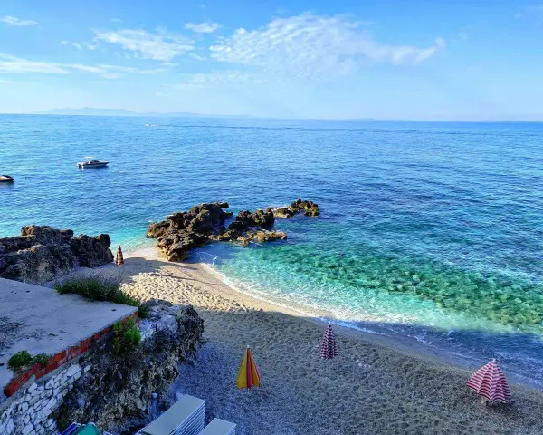 Albania, czerwiec: słoneczna pogoda nad krystalicznie czystym morzem, kamienista plaża z parasolami i łódkami na horyzoncie.