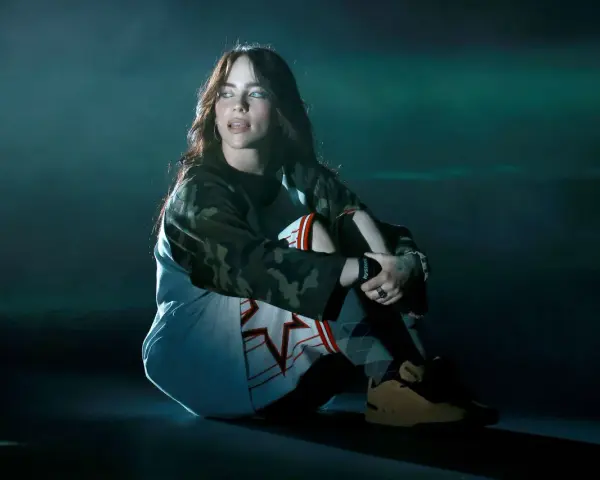 Billie Eilish siedzi na scenie, wpatrzona w dal. Jej stylizacja nawiązuje do estetyki jej albumu.