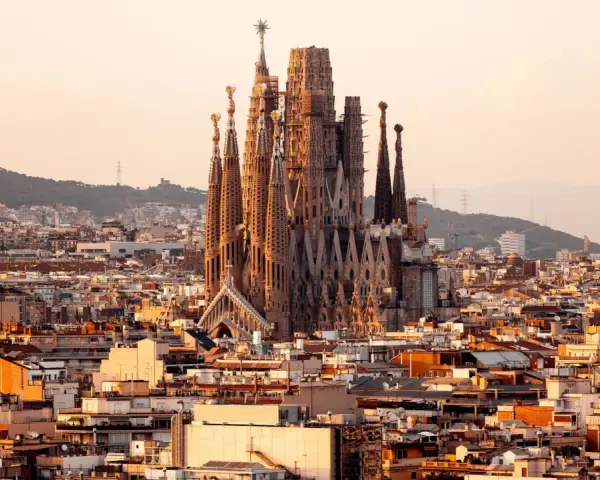 Vue panoramique de Barcelone au coucher du soleil, avec la Sagrada Familia au centre. Les travaux de la Sagrada Familia continuent, sa silhouette imposante se détachant sur le ciel orangé.