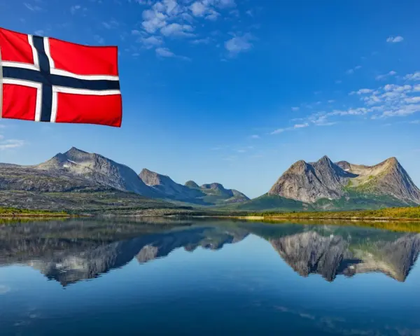 Norweska flaga powiewa nad jeziorem, odbijając się w wodzie. Czy do Norwegii potrzebny jest paszport? Piękne góry w tle.