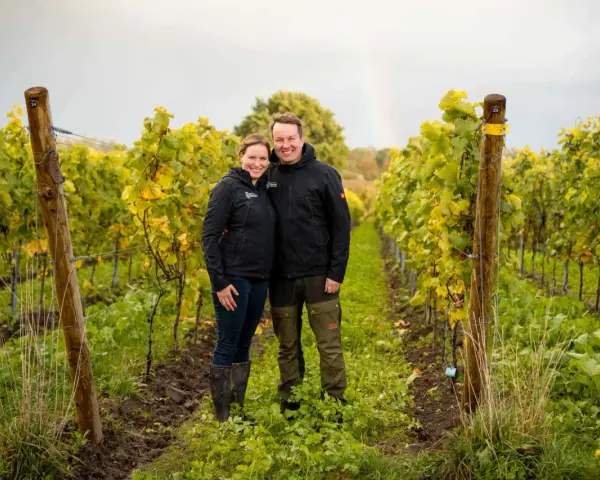 Ein Paar steht lächelnd in einem Weinberg in Schleswig-Holstein. Im Hintergrund ist ein Regenbogen zu sehen.