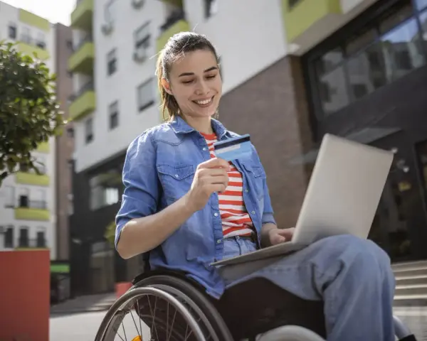 Uśmiechnięta kobieta na wózku inwalidzkim, korzystająca z laptopa i karty płatniczej, pokazuje, że stopnie niepełnosprawności nie ograniczają jej aktywności.