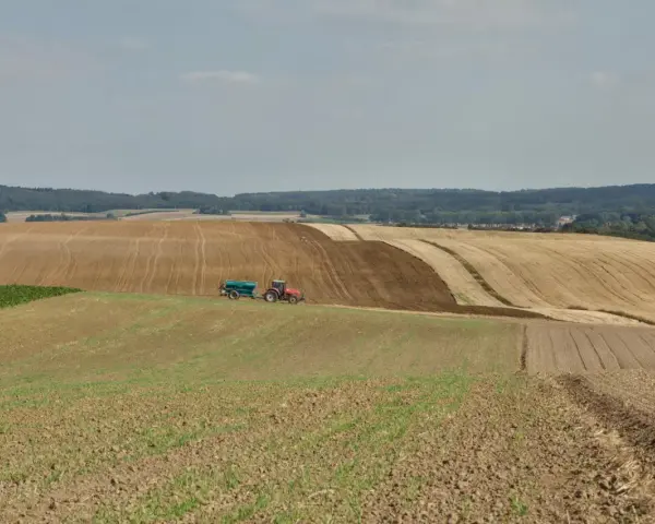 Czerwony traktor z przyczepą nawozi pole, przygotowując je do dalszej rekultywacji. W tle widać kolejne pola i las.