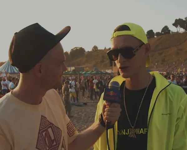 Dwóch mężczyzn na plaży, jeden z mikrofonem Red Bull, w tle tłum ludzi i scena.