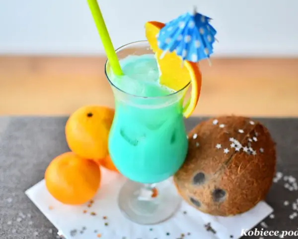 Orzeźwiający drink z sokiem ananasowym, ozdobiony plasterkiem pomarańczy i parasolką. Obok leżą pomarańcze i kokos.