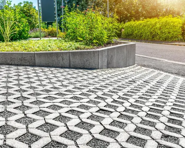Przestrzeń z ażurową nawierzchnią, która zapewnia minimalną powierzchnię biologicznie czynną, otoczona zielenią i betonowym murkiem.