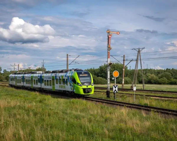 Zielony pociąg hybrydowy PKP jedzie po torach wśród zielonych pól. W tle las i chmury.