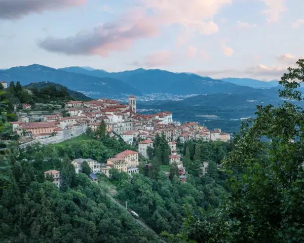 Borgo pittoresco su un colle, con case dai tetti rossi e un campanile, incastonato tra montagne e alberi. Via Francisca del Lucomagno, un incanto.