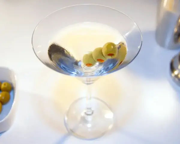 Elegancki drink z martini, ozdobiony trzema oliwkami, czeka na spróbowanie. Obok miseczka z oliwkami i shaker.