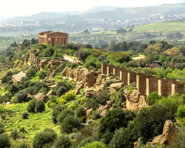 Ancient Greek temples stand proudly on a ridge in the Valley of the Temples, Sicily, surrounded by lush greenery and a distant town.