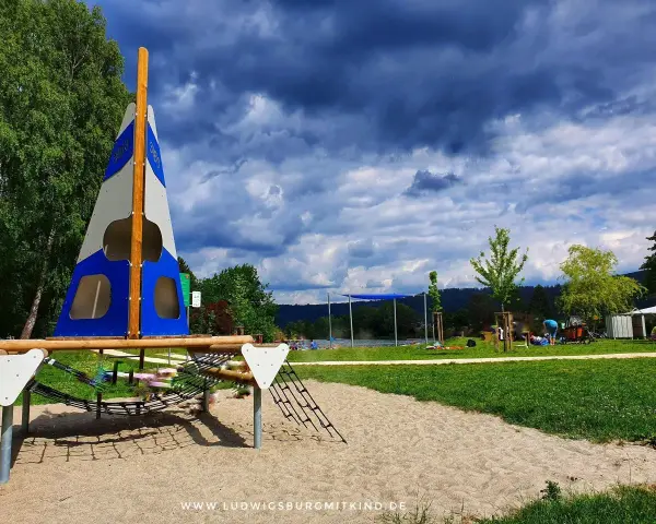 Spielplatz am Badesee Ludwigsburg mit Segel-Klettergerüst und Sandkasten. Im Hintergrund der See und Menschen.