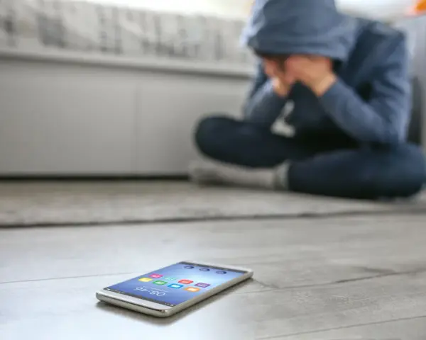 Osoba w kapturze siedzi na podłodze, ukrywając twarz w dłoniach. Obok leży telefon, symbolizujący izolację i dystymię.