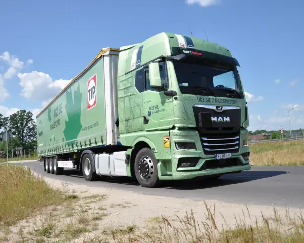 Zielona ciężarówka MAN TGX z naczepą TIP jedzie drogą. Na naczepie widnieje napis "Ucz się z najlepszymi!".