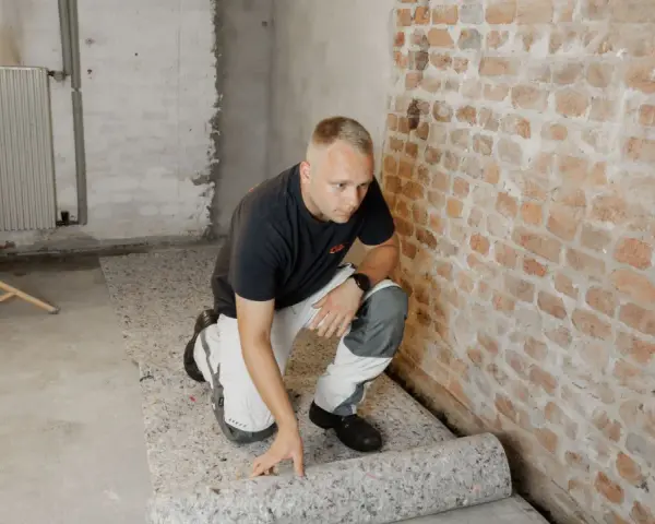 Mann rollt atmungsaktiven Bodenbelag im Keller aus. Die Ziegelwand und der Betonboden sind sichtbar.