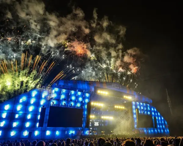 Nocny koncert z fajerwerkami nad sceną. Tłum ludzi podziwia widowisko, a nad nimi rozbłyskują kolorowe petardy.