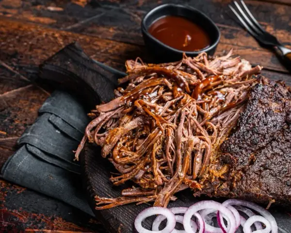 Soczysta, aromatyczna szarpana wołowina, polana sosem BBQ, podana z krążkami cebuli na drewnianej desce. Idealna na grilla.