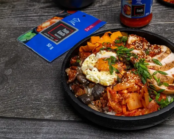 Pyszny Bibimbap przepis: ryż, kurczak, jajko sadzone, grzyby, marchewka i kimchi. Obok opakowanie pasty kimchi.