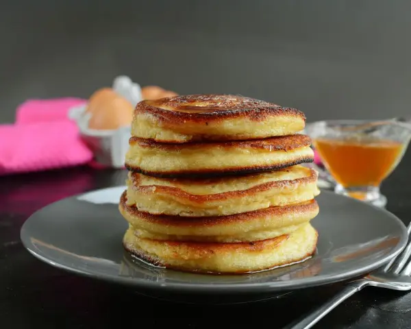 Pila di pancake senza latte, dorati e soffici, con sciroppo d'acero e uova sullo sfondo.