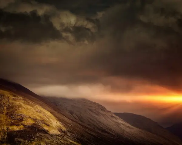 Dramatic sunburst pierces dark clouds over a rugged mountain range, a powerful display of the sovereignty of God.
