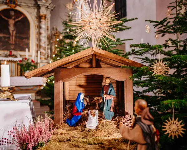 Weihnachtskrippe in einer Kirche, die die Geburt Jesu darstellt. Maria und Josef knien vor dem Kind, während im Hintergrund ein Weihnachtsbaum und Kerzen leuchten. So feiert man Weihnachten.